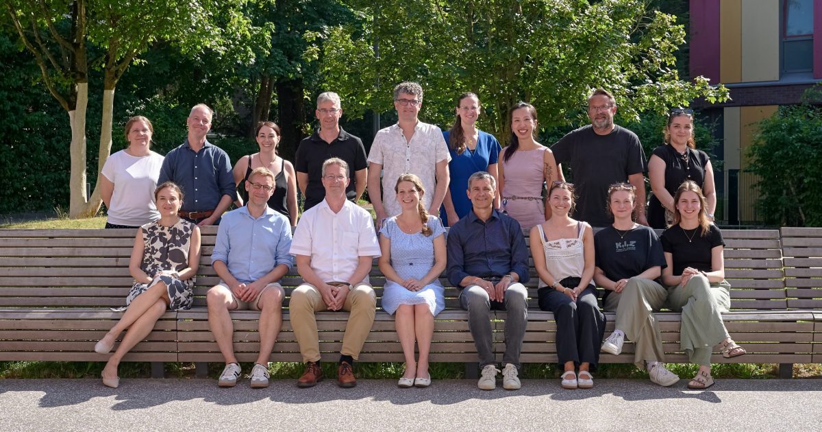 Group of adults posing outdoors on a long wooden bench, smiling for a team or class photo on a sunny day.