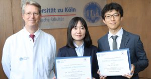 Three young professionals in formal attire hold certificates of appreciation, standing in front of a wooden wall with a university sign behind them.