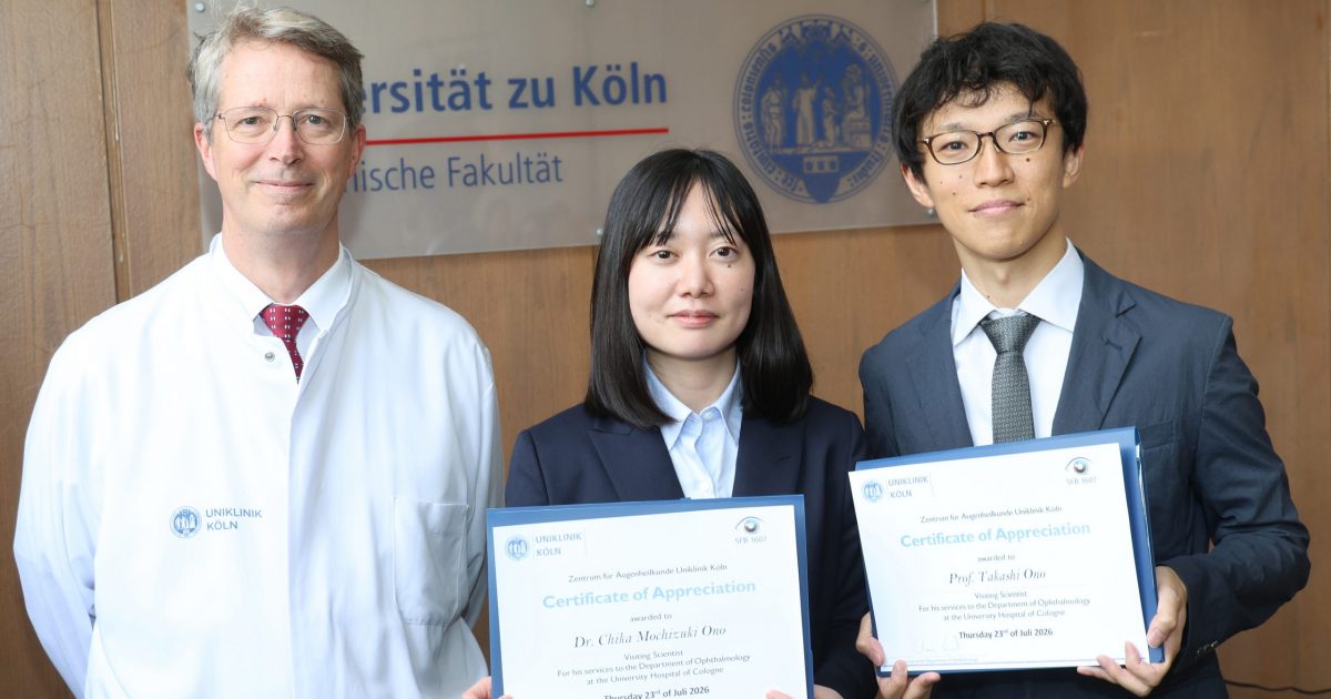 Three young professionals in formal attire hold certificates of appreciation, standing in front of a wooden wall with a university sign behind them.